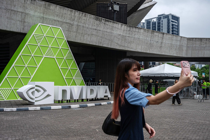 A woman takes a selfie in front of a large Nvidia sign with a green geometric design outside a conference venue