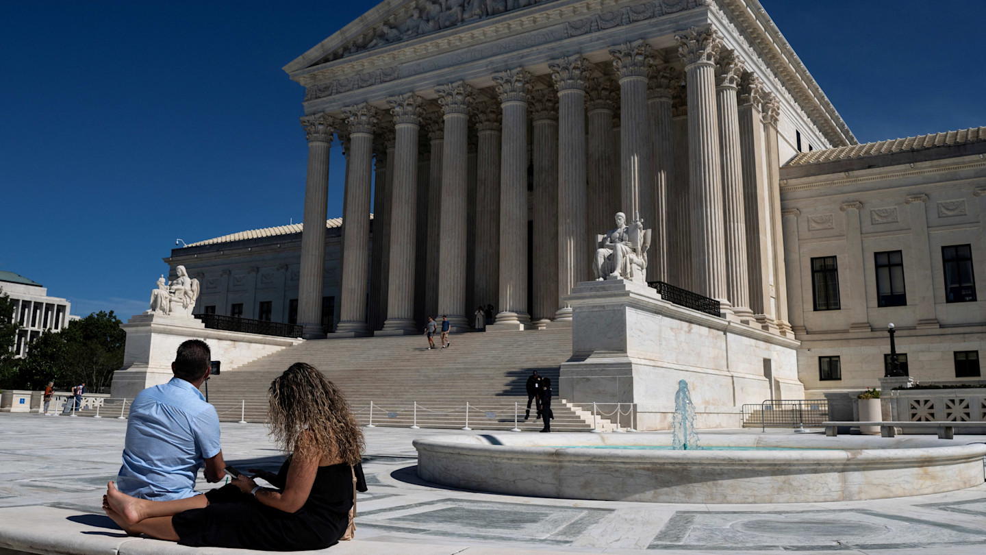 Two people sit beside a fountain outside the US Supreme Court building as others walk on the steps under a clear sky.