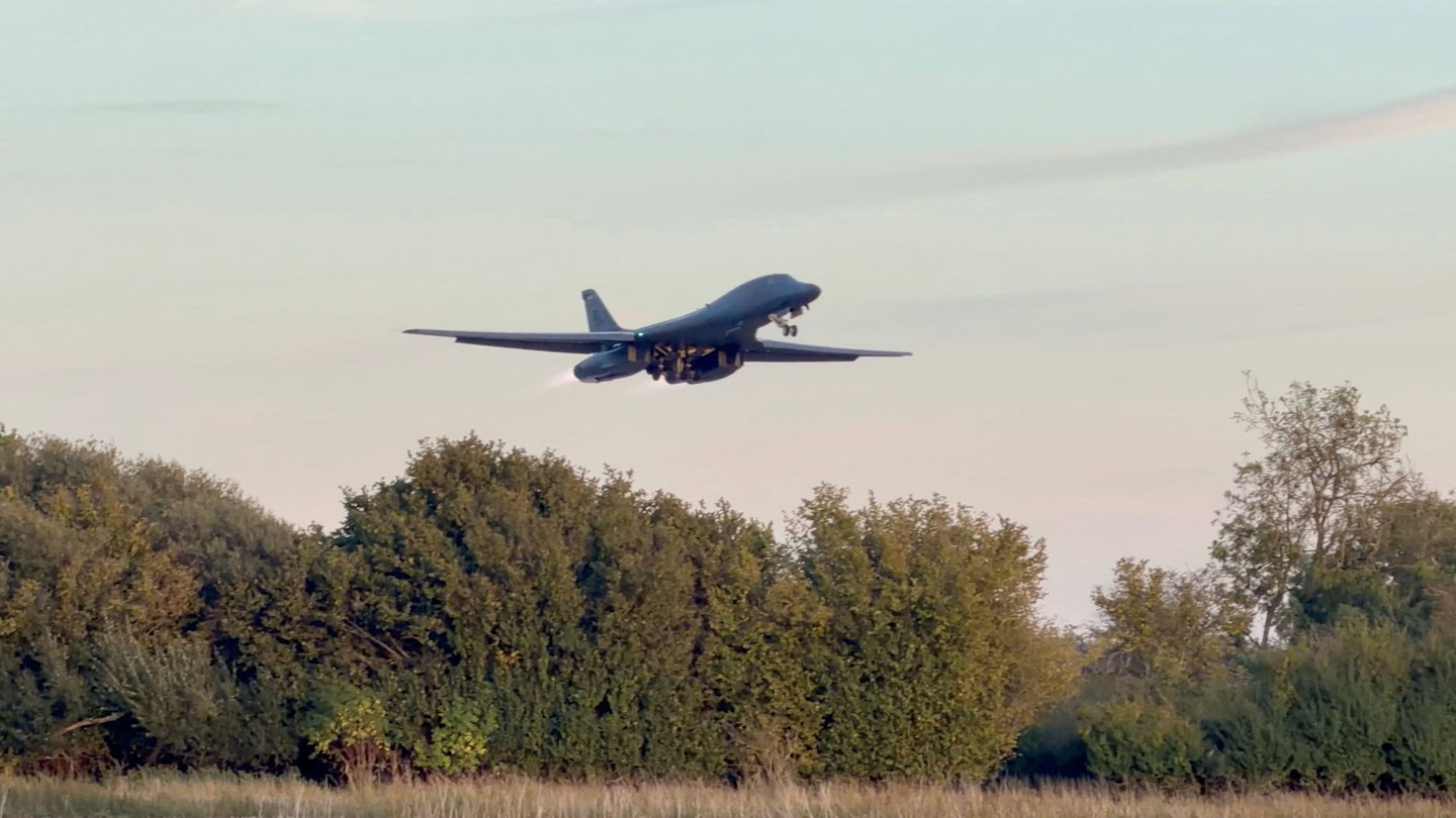 A B-1 bomber taking off above treetops, landing gear extended, at RAF Fairford airbase.