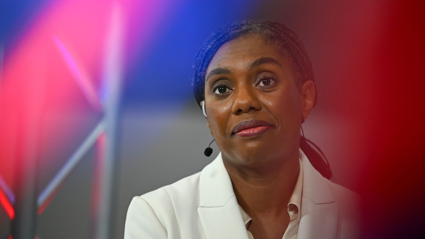 Kemi Badenoch wears a white blazer and headset microphone, with coloured stage lights blurred in the foreground.