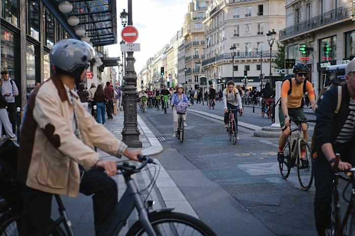 Several cyclists ride along a busy Rue de Rivoli in Paris, surrounded by pedestrians and city buildings on a clear day.