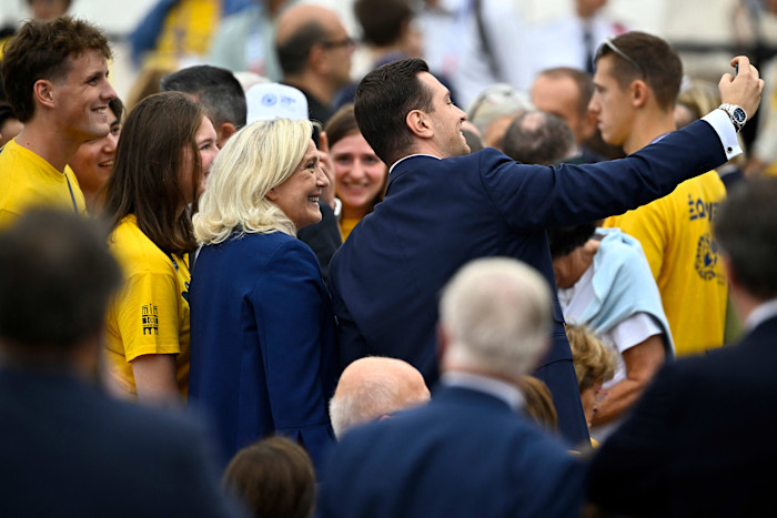 Marine Le Pen and Jordan Bardella smile as they take a selfie together among a crowd, including people in yellow shirts.