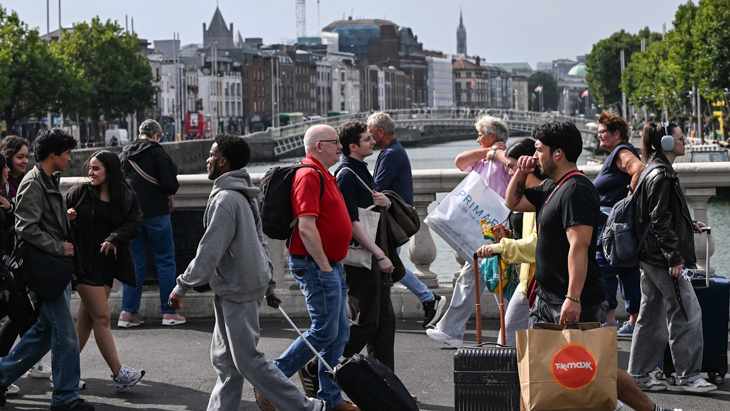 Pedestrians cross O’Connell Bridge in Dublin, many carrying shopping bags and luggage, with the River Liffey and city buildings in the background.