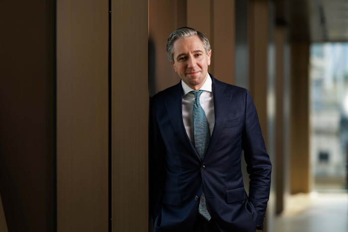Simon Harris stands indoors, leaning against a wall and looking at the camera, wearing a navy suit and teal tie.