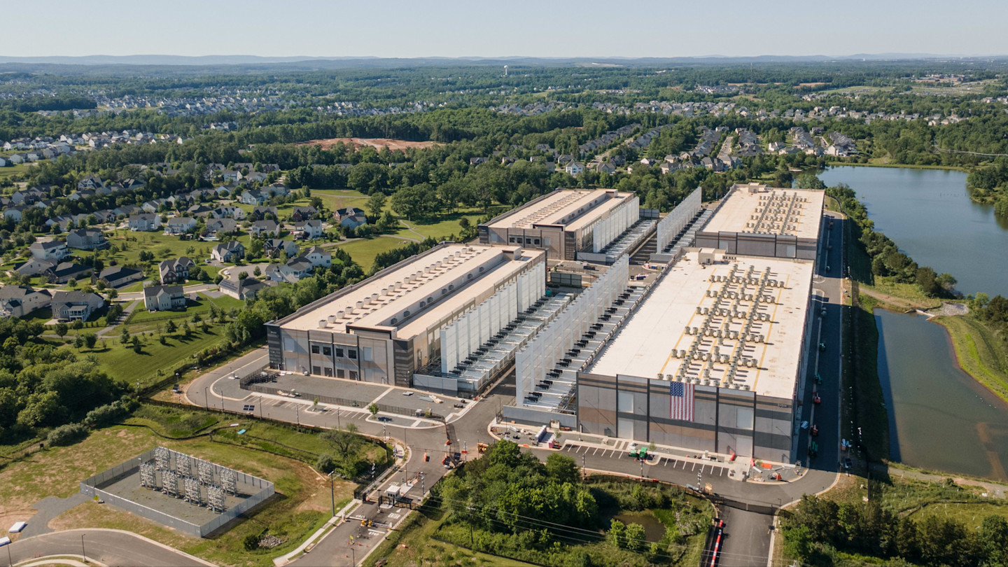 Aerial view of a large Amazon Web Services data centre complex surrounded by residential neighbourhoods and trees.