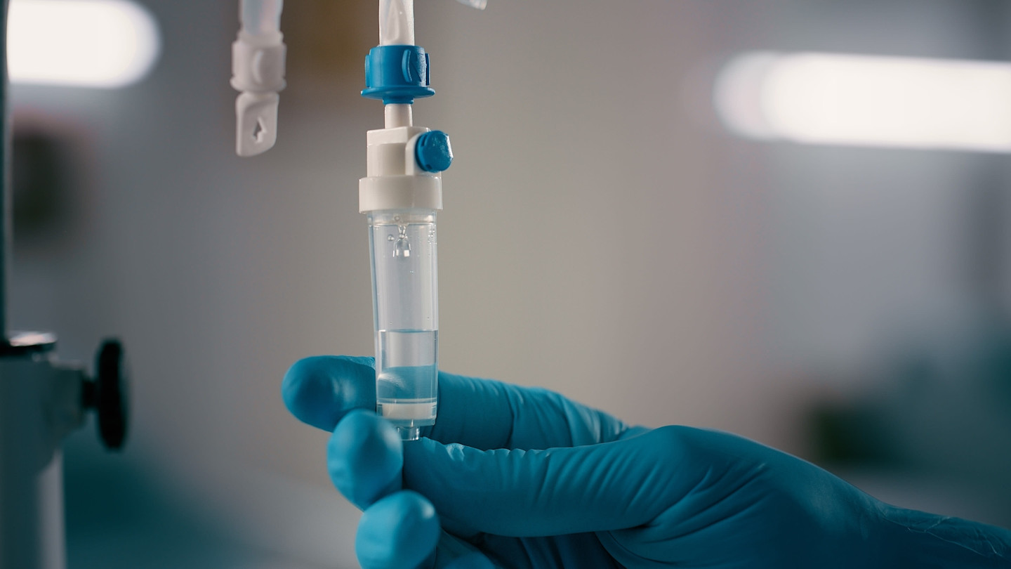 A gloved hand adjusts an IV drip chamber filled with clear fluid in a clinical setting.