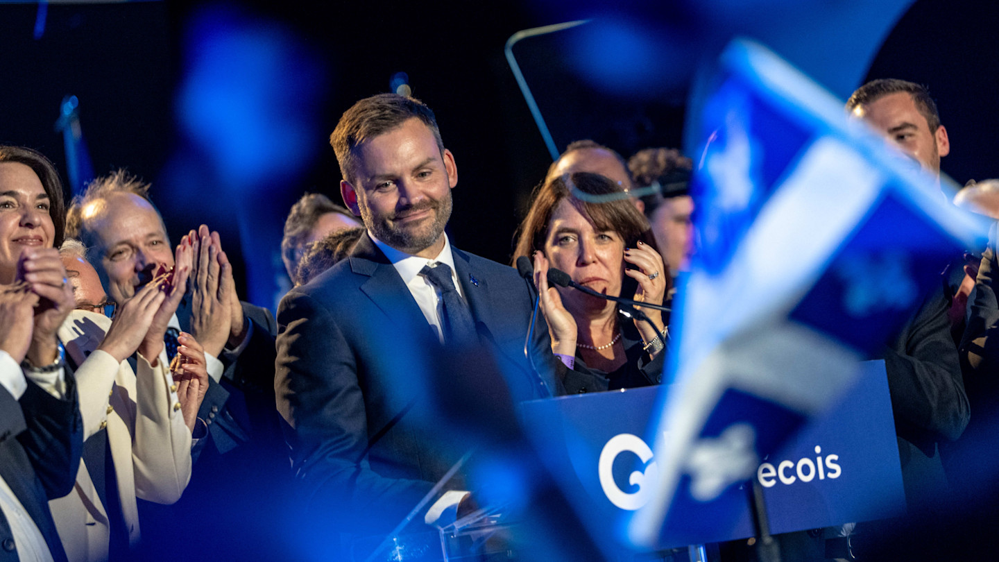 Paul St-Pierre Plamondon stands at a podium surrounded by supporters, smiling as people applaud and wave a Quebec flag in the foreground.