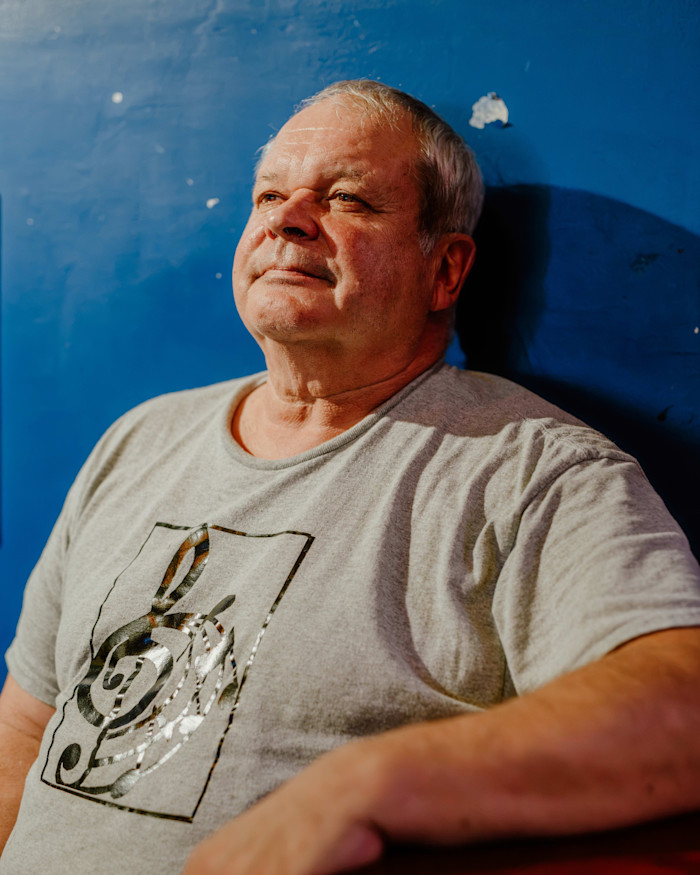 Clem Jaskot Jr. sits against a blue wall, wearing a gray t-shirt with a musical note graphic.