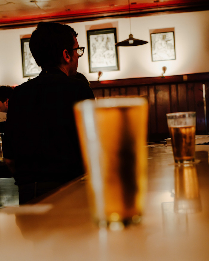 A person sits at the bar with two glasses of beer in the foreground, inside Rainbow Club in Chicago’s Wicker Park.