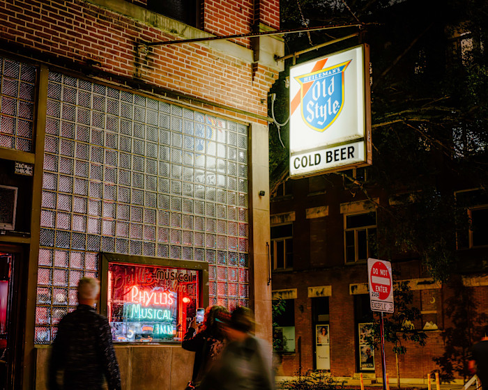 Neon sign for Phyllis Musical Inn glows in a glass-block window beneath a "Heileman's Old Style Cold Beer" sign at night, with blurred people passing by.