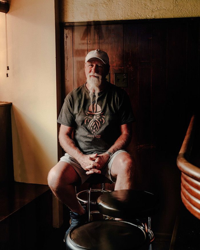 Scott Martin sits on a barstool inside Simon’s Tavern, hands clasped, wearing a t-shirt and white cap.