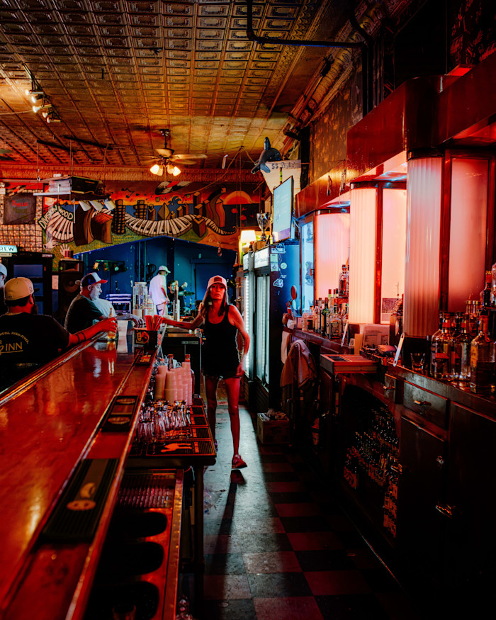 A bartender walks behind the bar at Phyllis Musical Inn, with colorful lights and a mural decorating the interior.