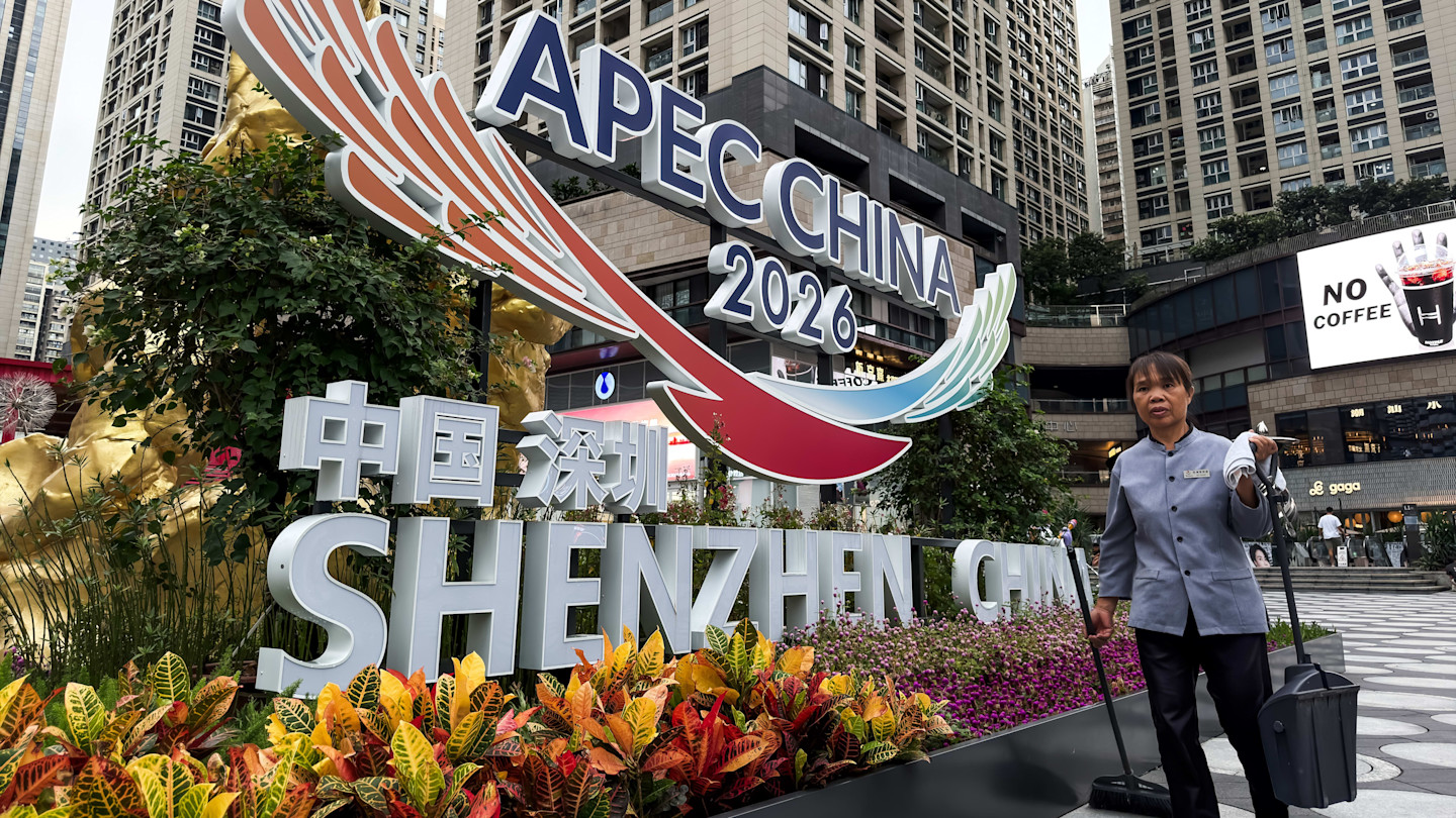 A cleaner holding cleaning tools stands beside a large Apec China 2026 promotional display in front of high-rise buildings and shops in Shenzhen.
