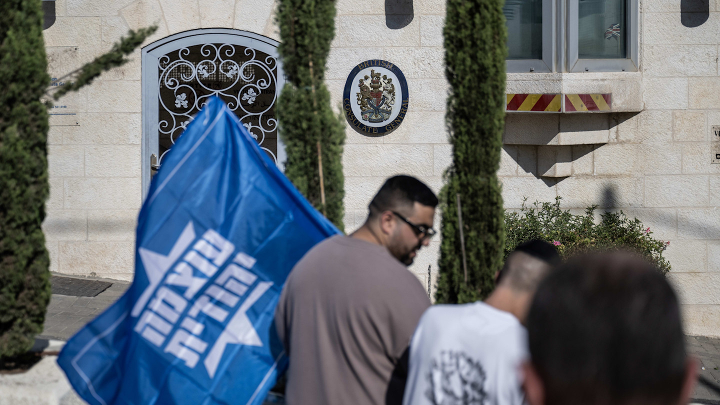 A protester holds a blue flag with Hebrew text outside the British Consulate General in Jerusalem. The consulate's emblem is visible on the building.