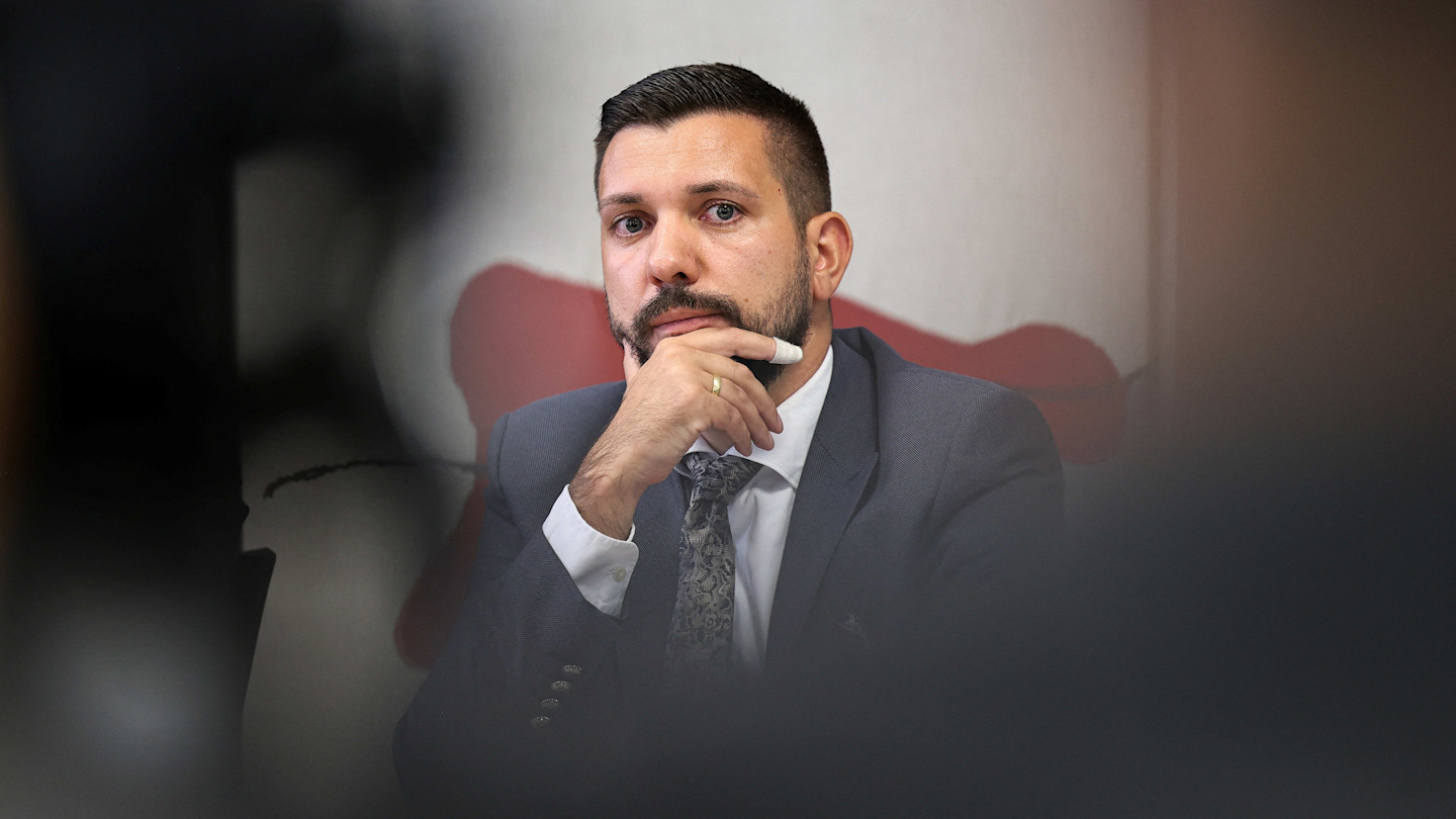 Tobias Rausch sits at a press conference table, resting his chin on his hand and looking forward thoughtfully.