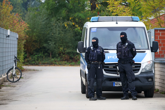 Two police officers in tactical gear and balaclavas stand in front of a police van near the entrance to a residence.