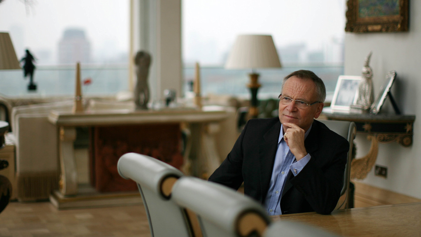 Jeffrey Archer sitting at a table in a well-lit room with large windows, appearing thoughtful.