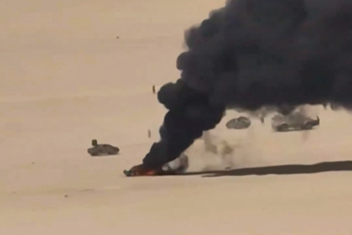 Aerial view of a burning vehicle emitting thick black smoke on a desert landscape, with several other vehicles nearby.