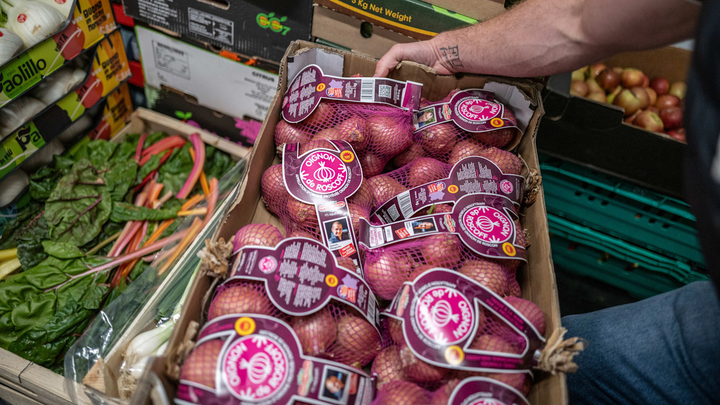 A person holds a box of packaged Roscoff onions among crates of assorted fresh produce at a market.