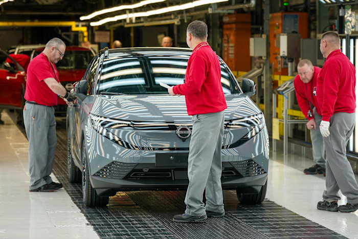 Four workers in red shirts and grey trousers inspect a grey Nissan Leaf electric vehicle on the factory floor.