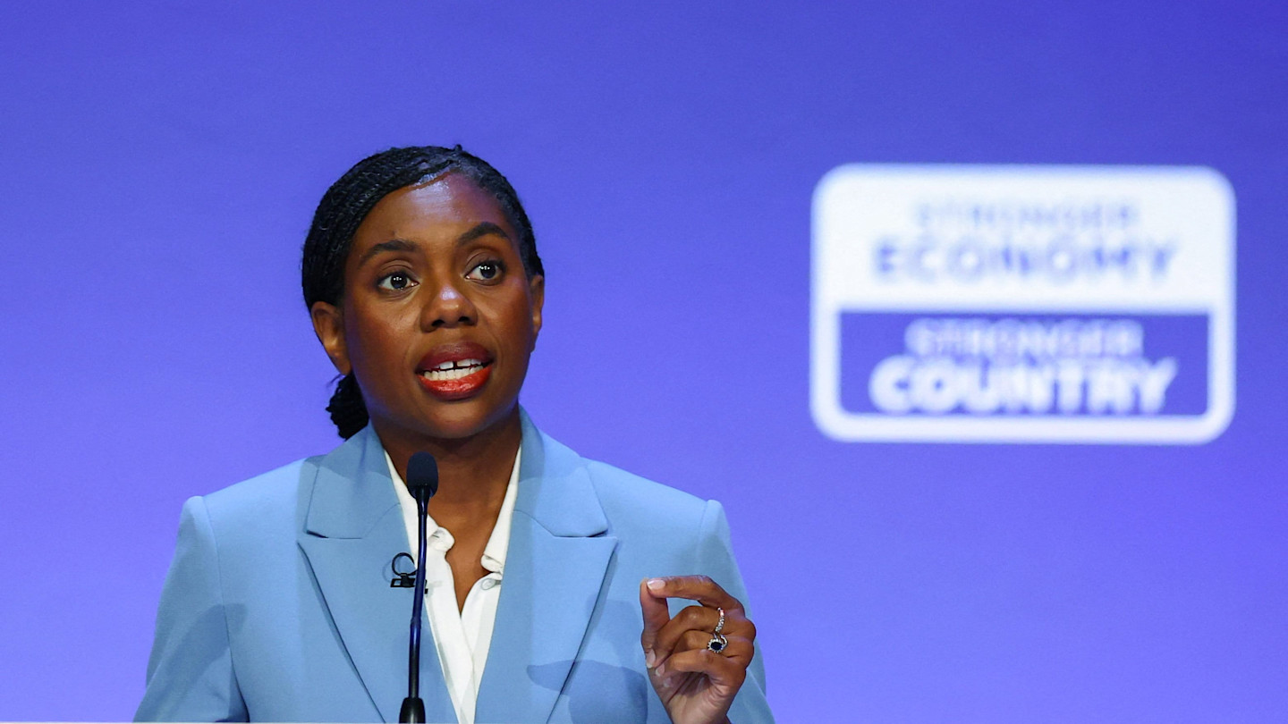 Kemi Badenoch speaks at a podium against a blue backdrop with a blurred sign reading "Stronger Economy, Stronger Country."