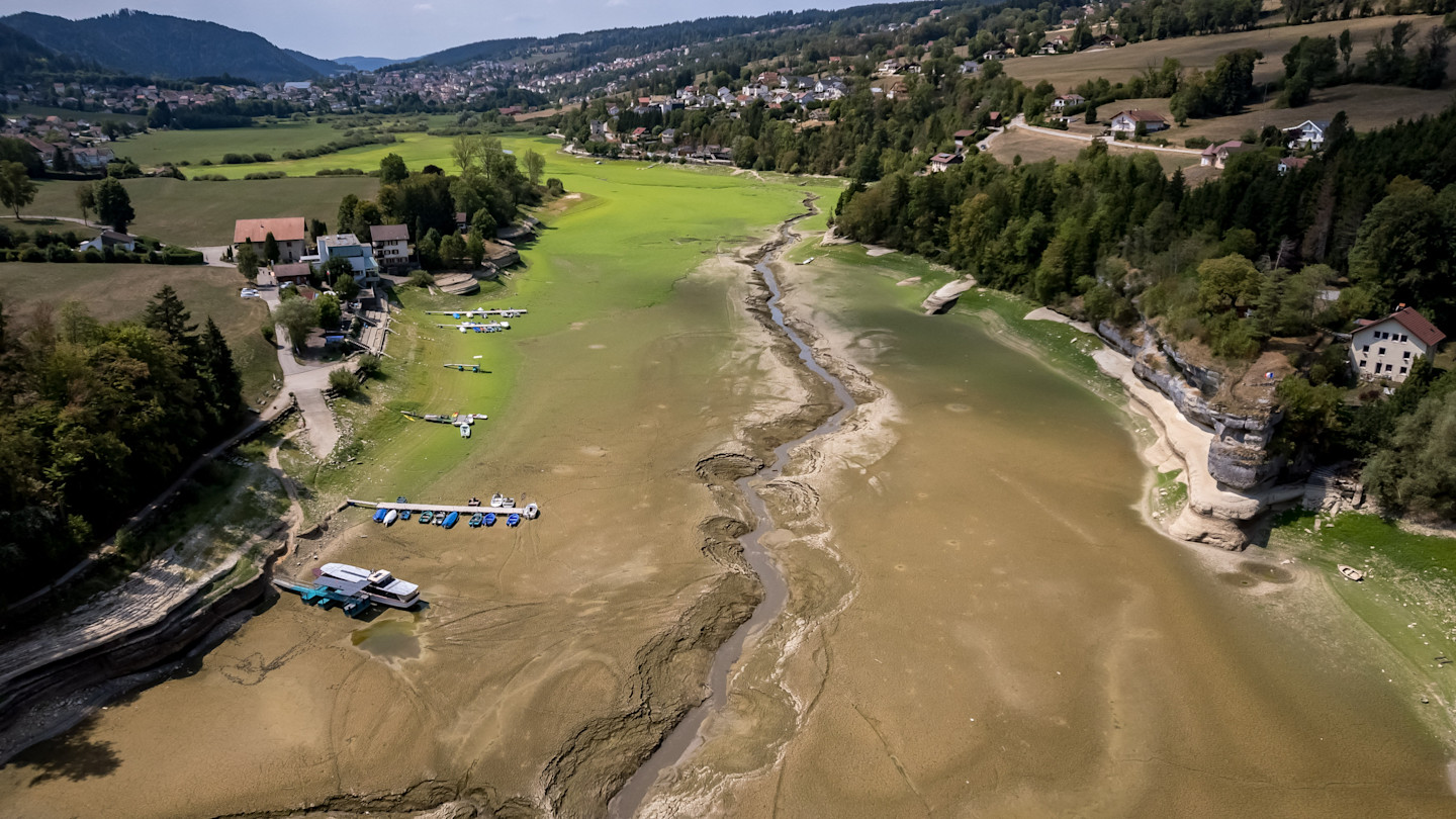Aerial view of stranded boats on the dry, cracked bed of Brenets Lake, with only a narrow stream of water remaining.
