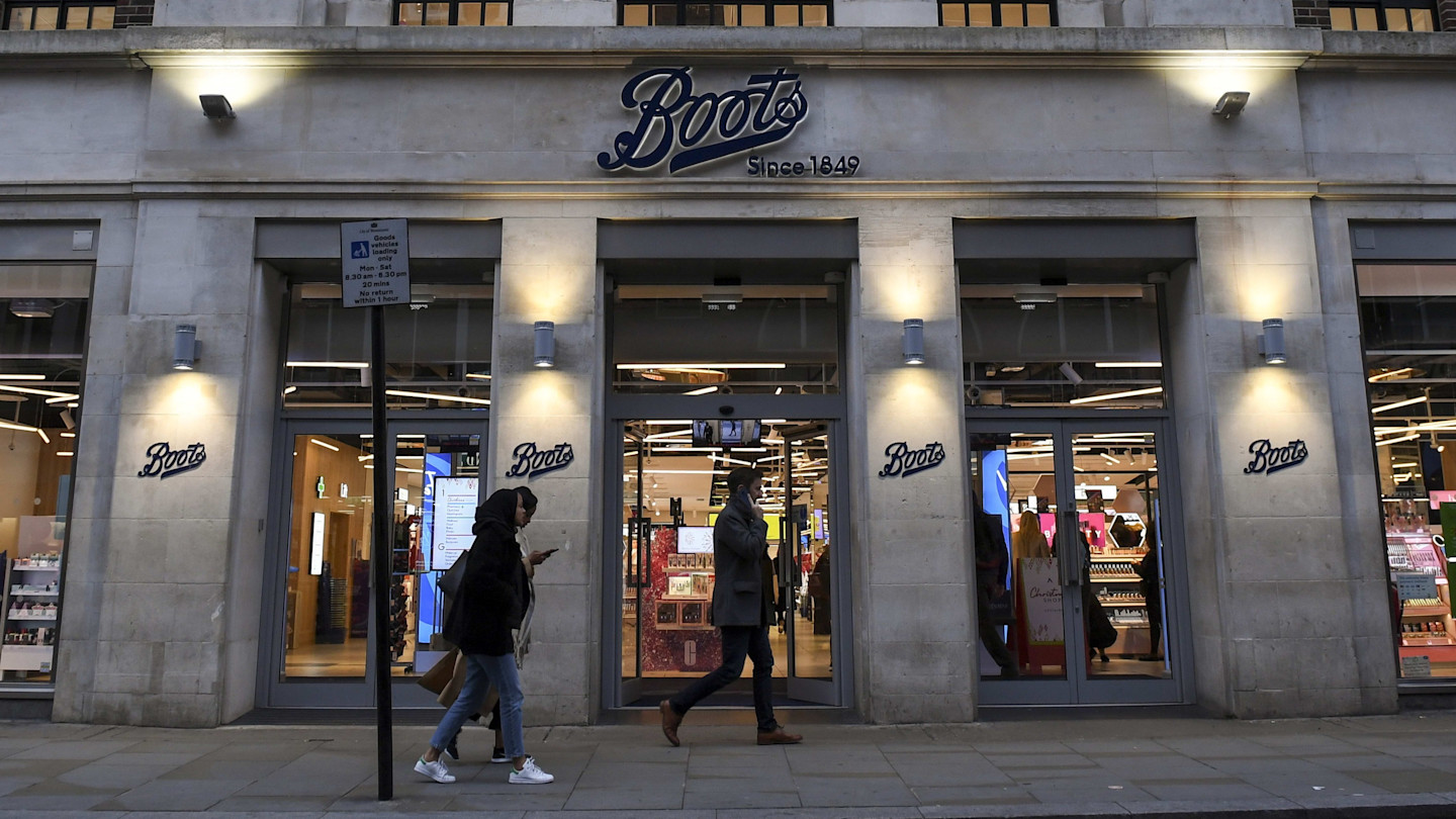 Pedestrians walk past the entrance of a Boots chemist store in London, with illuminated signs above and beside the doors.