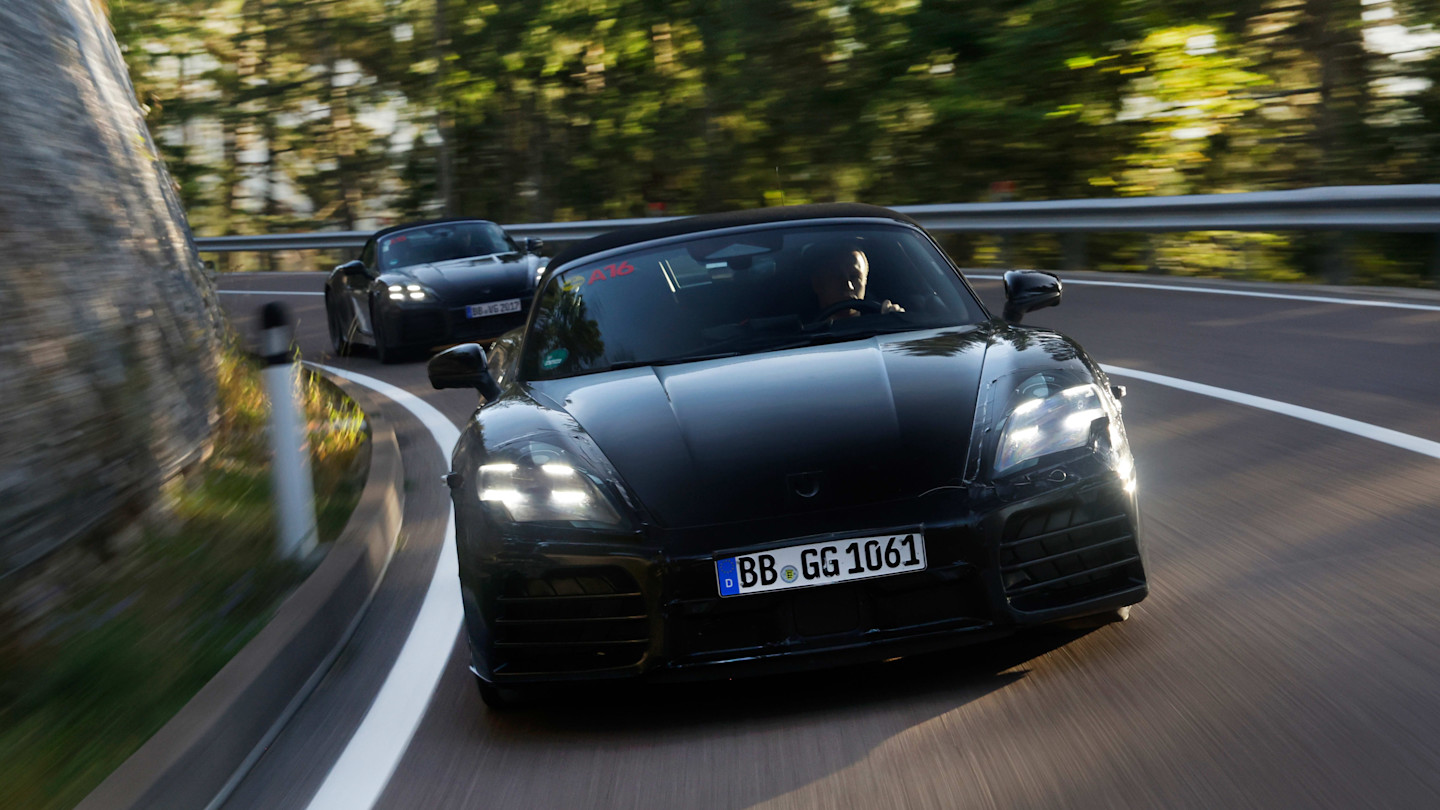 Two black prototype Porsche 718 Boxster sports cars driving on a winding road, with the front car in sharp focus.