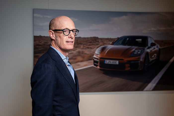 Michael Leiters stands indoors in front of a large photograph of a Porsche driving on an open road.