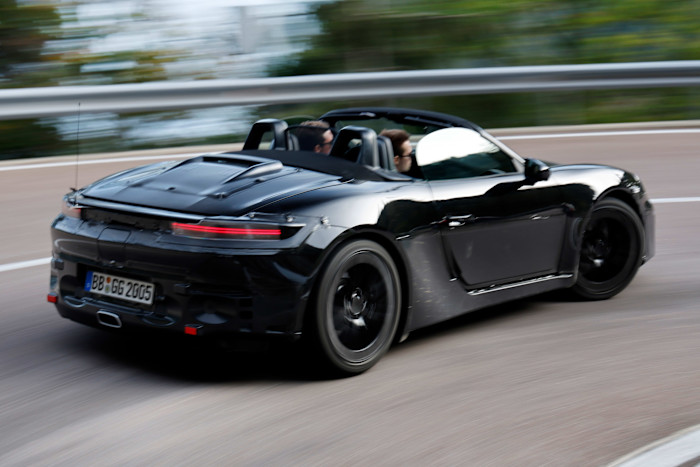 A black prototype Porsche 718 Boxster convertible is driven on a curving road, with two people visible inside.