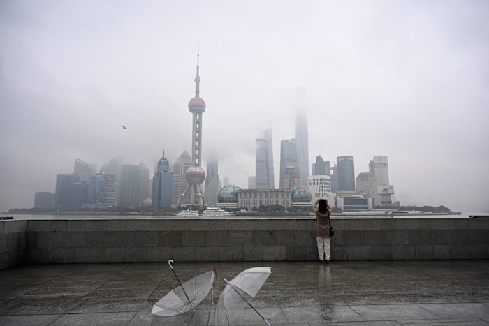 A woman takes a photo of Shanghai’s Lujiazui skyline on a foggy day, with two transparent umbrellas on the wet promenade in the foreground.