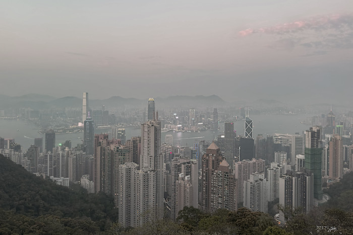 Hong Kong skyline at dusk with high-rise buildings emerging from mist, harbour visible, and sky tinged pink with soft clouds