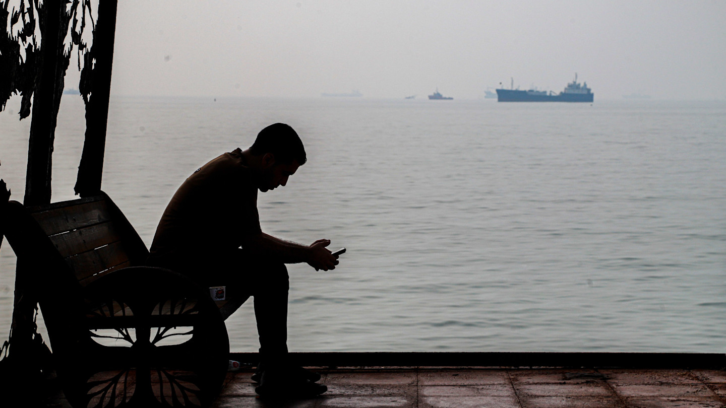 A man sits on a bench at a waterfront shelter, looking at his cell phone, as several commercial ships are anchored offshore.