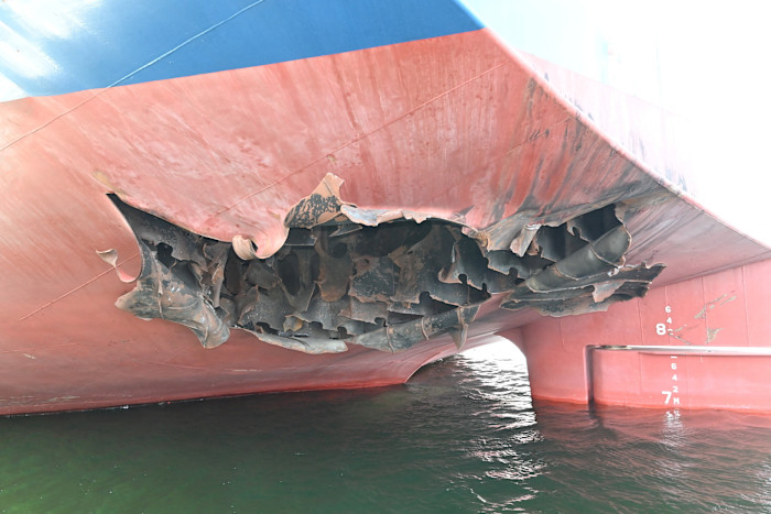 Hull of a bulk carrier operated by HMM with a large, jagged hole and twisted metal at the stern above the waterline.