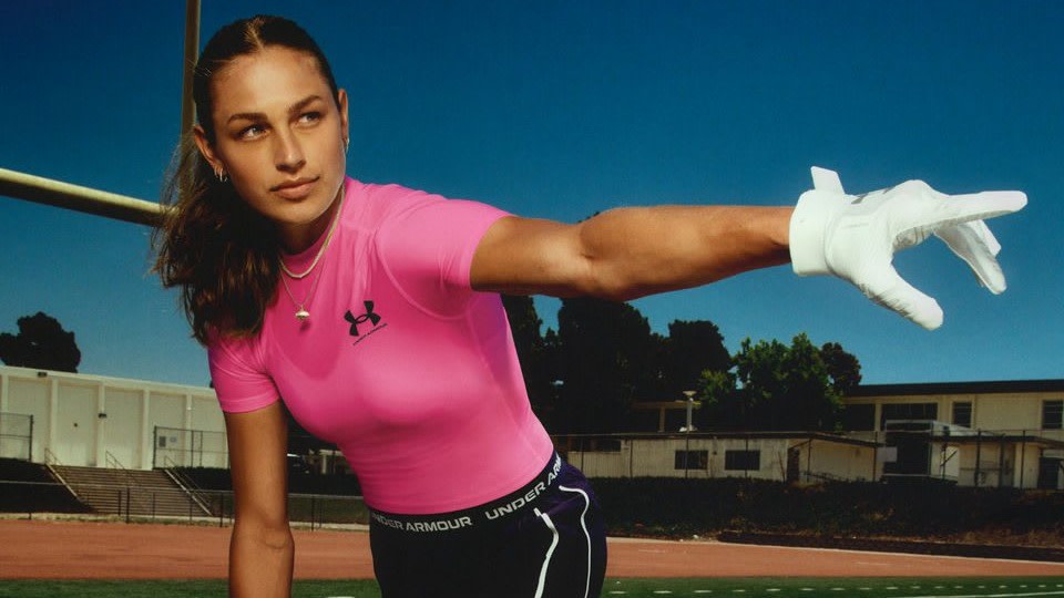 A person in a bright pink Under Armour shirt and white glove extends their arm on an outdoor sports field.