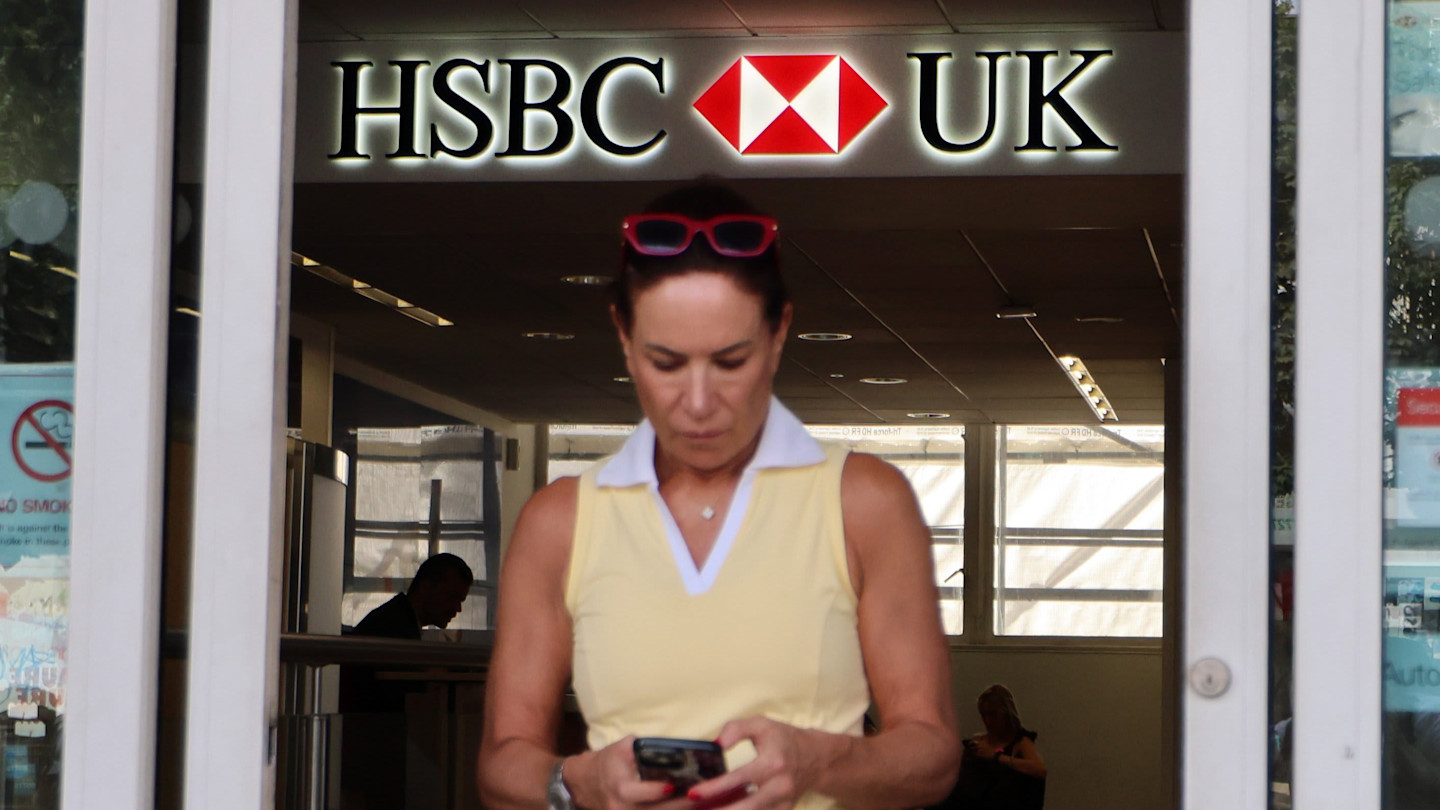 A woman looks down at her phone while exiting an HSBC UK bank branch in London. The HSBC logo is visible above her.