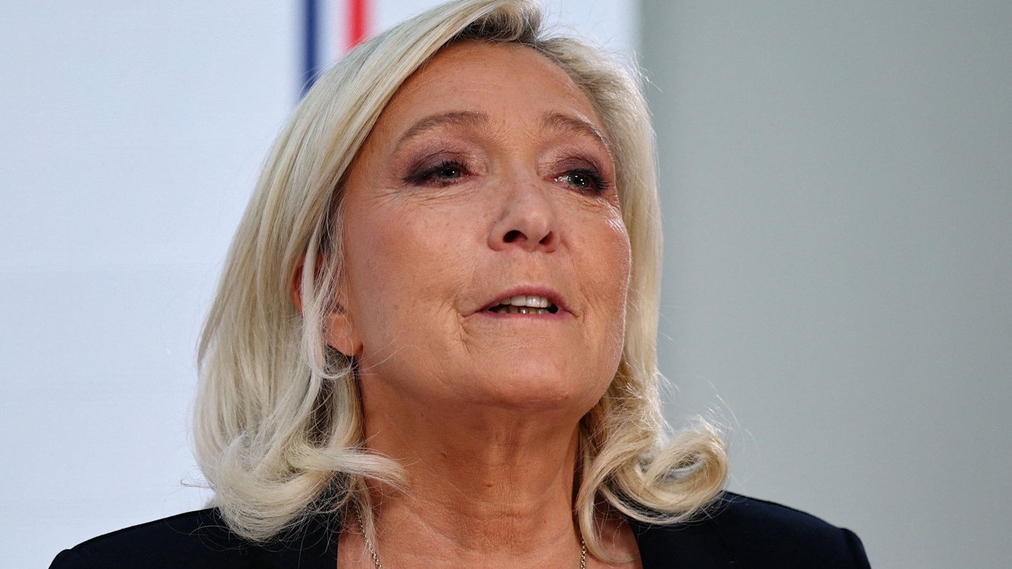 Marine Le Pen speaks at a press conference, with part of a French flag visible in the blurred background.