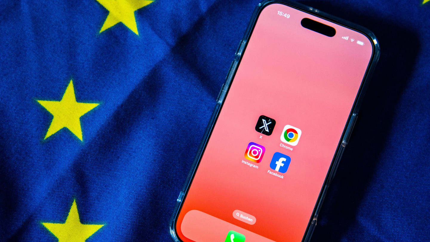 A smartphone displaying the app icons for X, Chrome, Instagram and Facebook rests on an EU flag