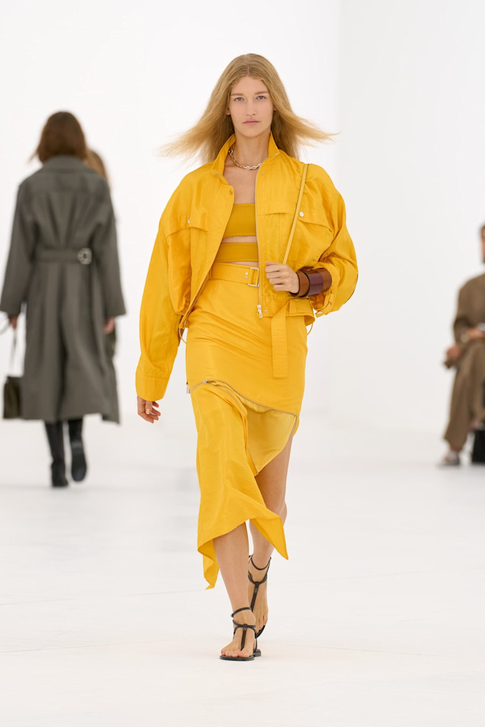 A model walks the runway in a mustard-yellow cropped jacket, bandeau top and asymmetric belted skirt, paired with black sandals.
