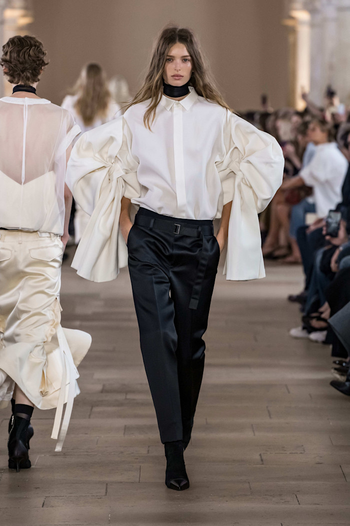 A model walks the runway in a white shirt with dramatically gathered sleeves, paired with belted black trousers and a black neckband.