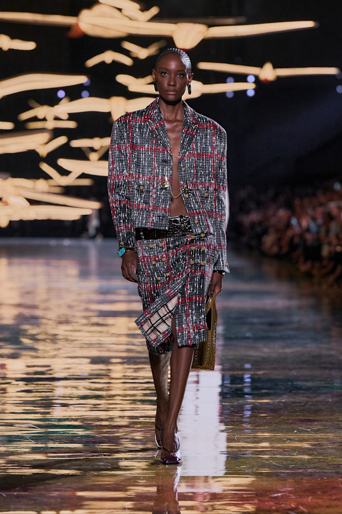 A model walks the runway in a grey and red checked cropped jacket and matching knee-length skirt, carrying a metallic bag.