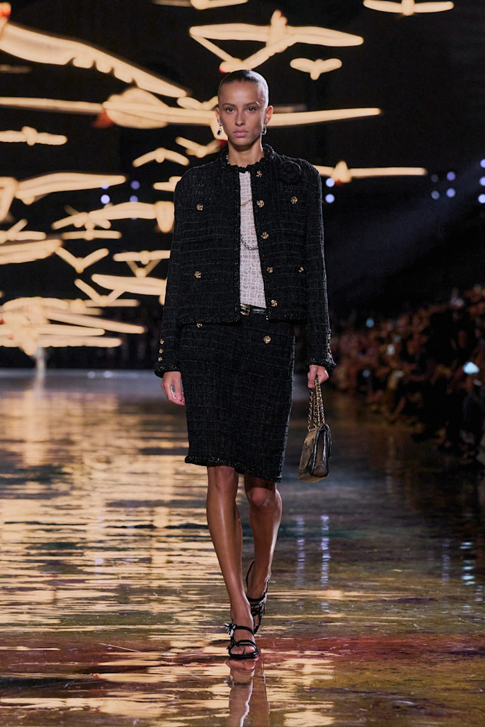 A model walks the runway in a black tweed jacket and matching knee-length skirt with gold buttons, carrying a small chain-strap bag.
