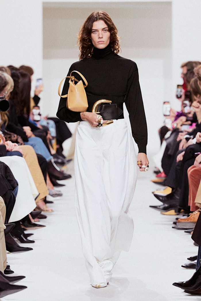 A model walks the runway in a black roll-neck top and voluminous white trousers, with an oversized black belt and tan handbag.
