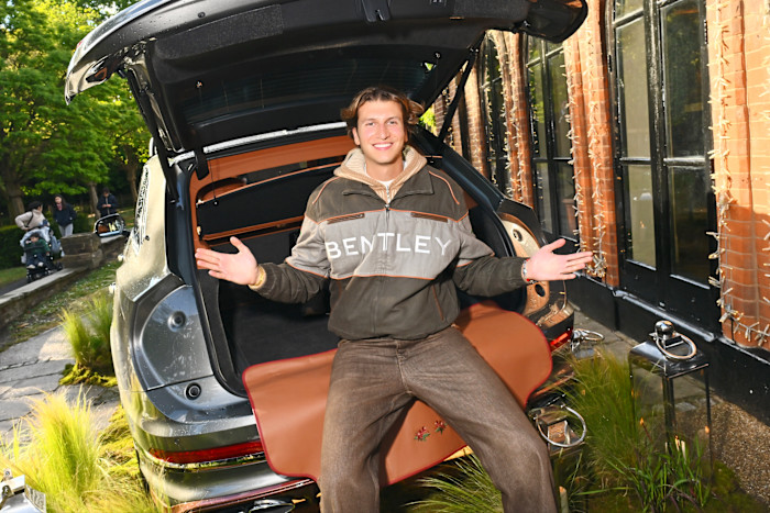 Gstaad Guy sits in the open trunk of a Bentley Bentayga, smiling with arms outstretched at an outdoor event.