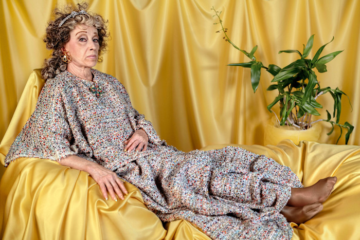 Cindy Sherman reclines on a yellow satin-draped surface in a textured multicoloured gown, with a potted plant in the background.