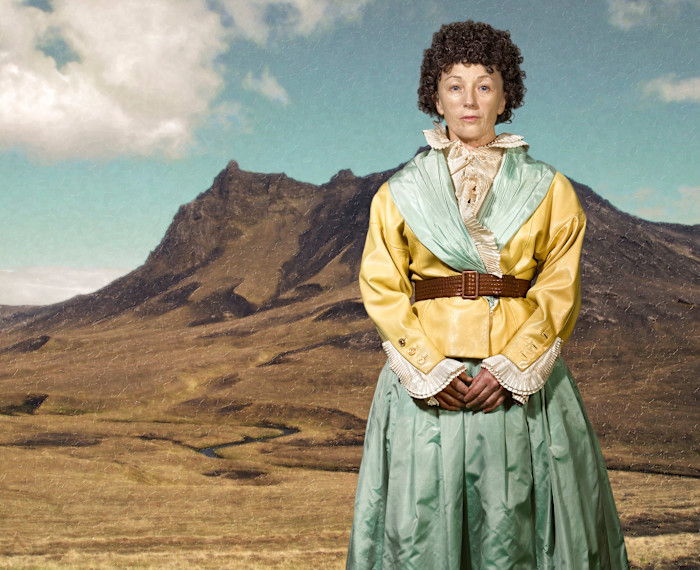 Cindy Sherman stands in front of a mountainous landscape, wearing a yellow and green period costume with a ruffled collar.