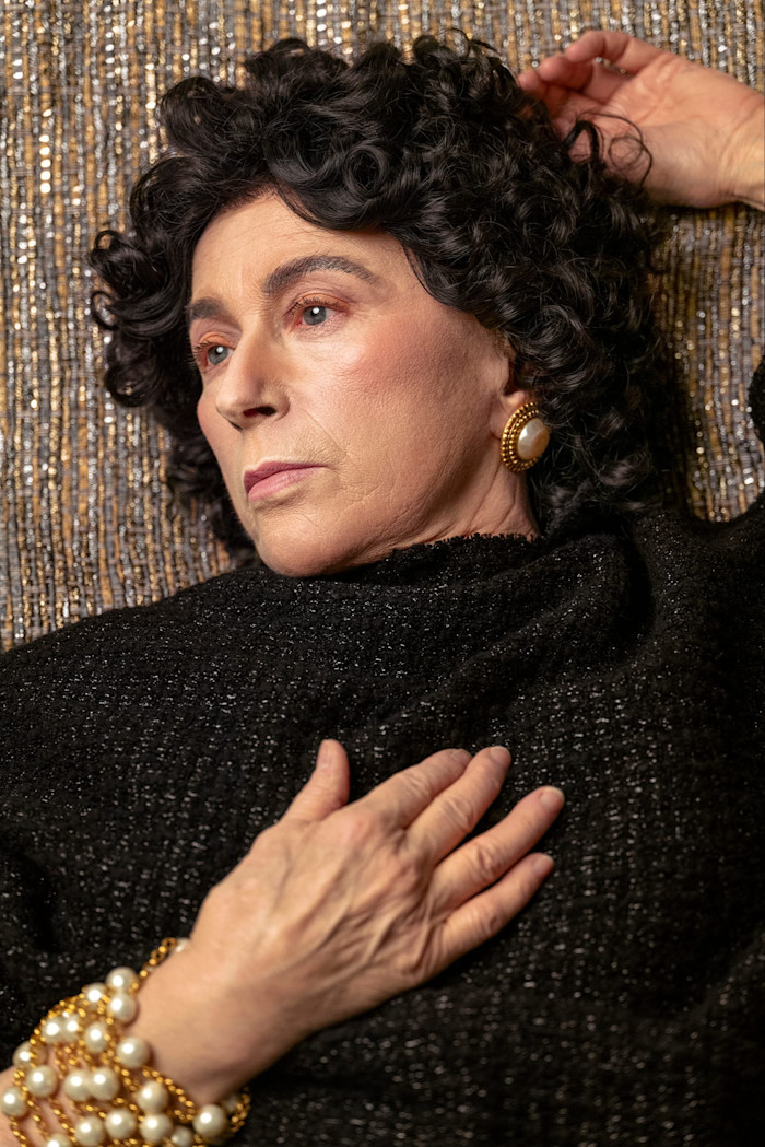 Cindy Sherman poses with a curly wig, pearl jewelry, and black clothing, reclining against a textured, metallic background.