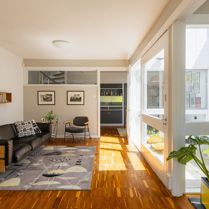 A bright living room with parquet flooring, a black leather sofa and modern furnishings. Sunlight streams in through large windows and glass doors. On the market with The Modern House.