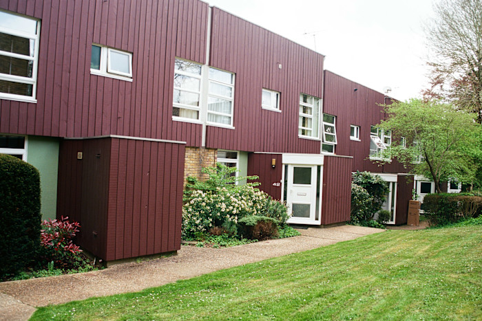 A row of modernist terraced houses with vertical maroon wood cladding, large windows and garden areas in front.