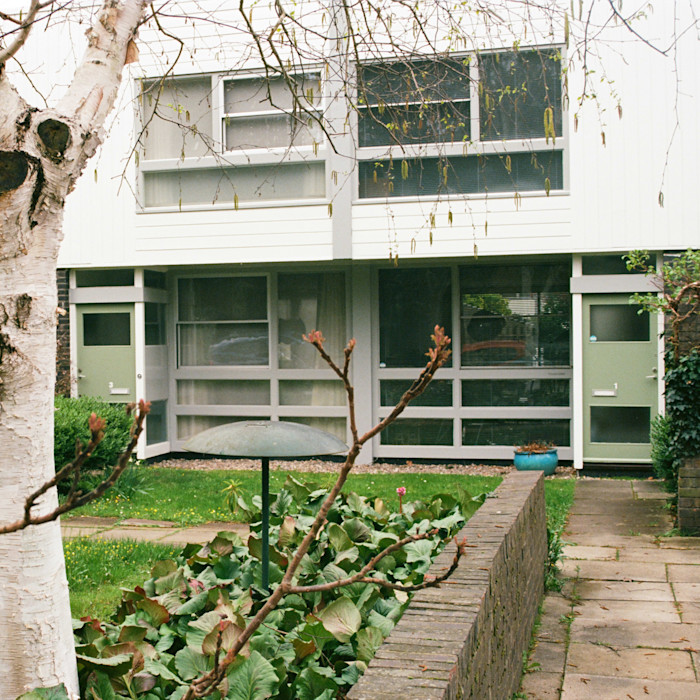 A two-story modernist townhouse with large windows and green doors, set behind a small garden and brick path.